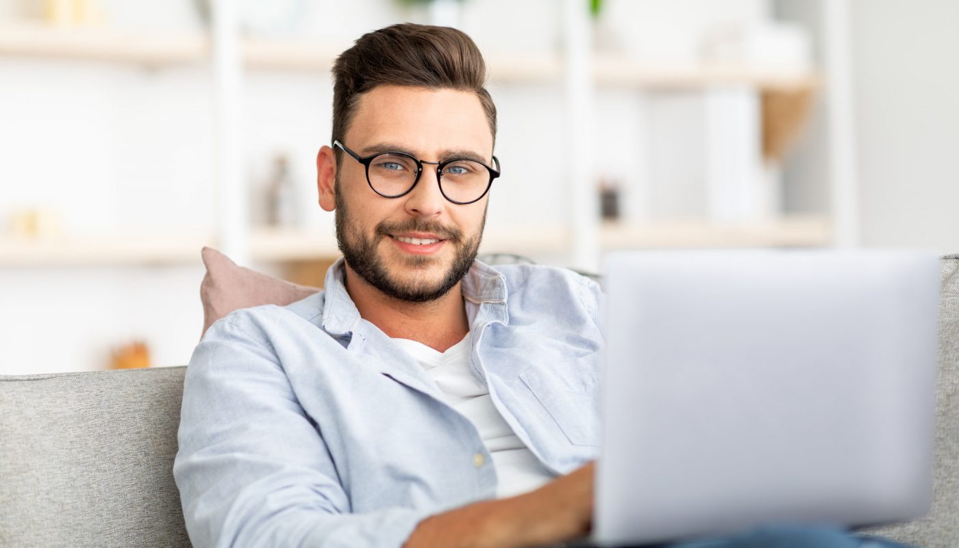 Handsome guy with glasses using laptop at home, sitting on sofa, working from home, typing emails or