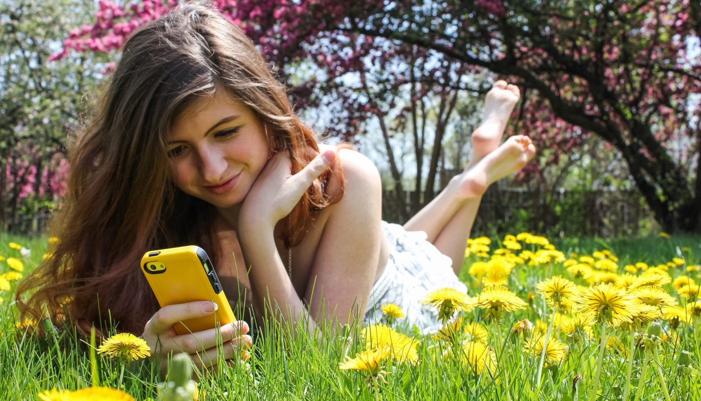 Young happy woman lying in grass and feet up taking photos of yellow dandelions using mobile phone